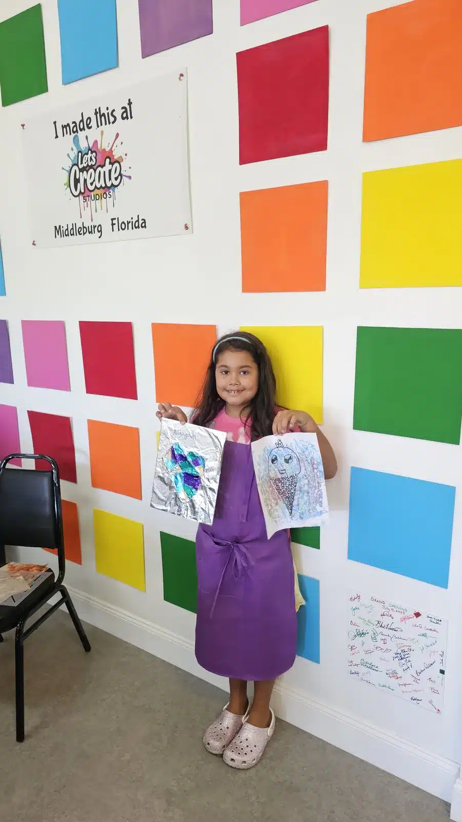 Girl showing her art projects in front of the 'I made this at Let's Create Studios' wall in Middleburg