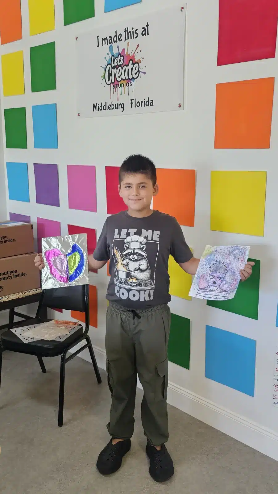 Boy holding his finished art projects in front of the colorful wall at Let's Create Studios, Middleburg
