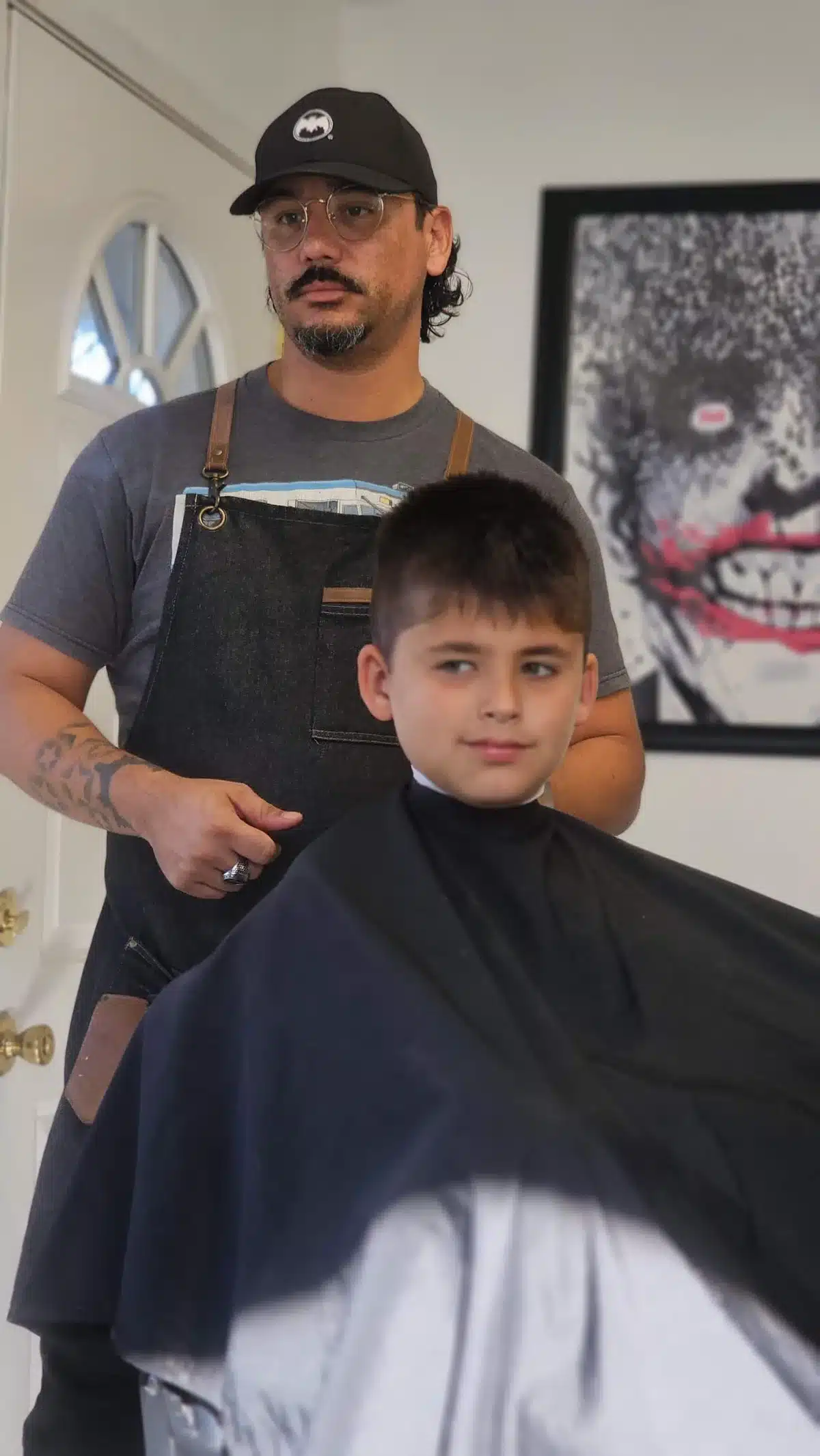 Portrait of Jay the barber in a black cap and denim apron standing behind a young client